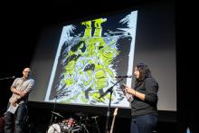 A man and a woman stand before microphones in front of a screen with a graphic novel image on it