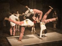 Three young men caught mid cartwheel and flip while other circus-like performers mill around on stage