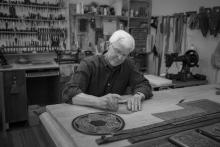 a gray-haired man sitting in a workshop working on a piece of leather