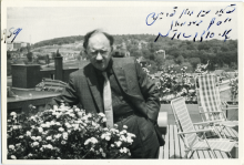 Black and white photo of a man wearing glasses and a suit standing on a balcony