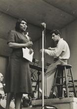 A woman in 1940s dress sings before a microphone while a man behind her conducts unseen musicians