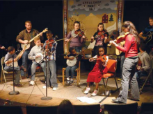 a racially diverse group of young people playing fiddles and banjos on a low stage