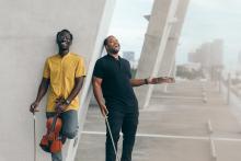 Two men stand against a pillar and laugh while holding violins