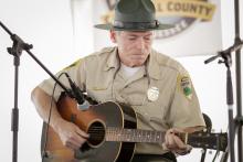 Bob Fulcher in his state parks uniform sits on a chair playing the guitar