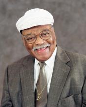 A man in a white hat, with glasses and a mustache smiles, wearing a small trumpet charm on a necklace.