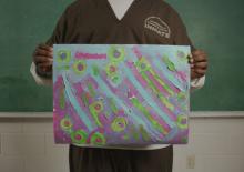 A young man in prison garb holds up a brightly colored painting that he made