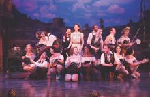 an ensemble of actors posed in a group on a stage holding various pots and pans and other implements