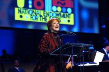 A woman with short red hair and a bright smile stands at a podium and microphone with a colorful sign in the background.