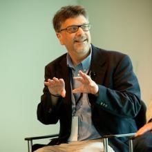 a bearded white man with glasses seated in a chair gesturing with his hands