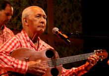 A man in a red checkered shirt plays the guitar