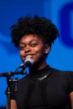 headshot of a young African American woman with her hair in an updo speaking into a microphone