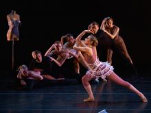 A female dancer in profile leans toward the left with her hand bent on her head while several dancers behind her mimic the move