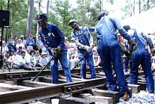 A group of men in work overalls demonstrate gandy dancing on replica railroad tracks