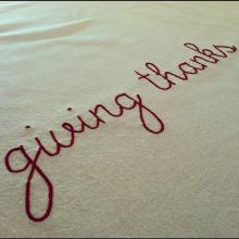 The phrase "giving thanks" embroidered in red thread on a white background