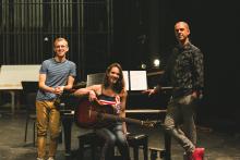 A young woman sits on a piano bench holding a guitar while two men stand casually leaning on the piano