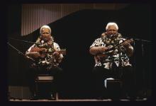 Two men on a stage playing ukuleles and singing.
