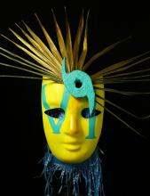 A yellow mask with "VI" painted on it in teal with gold palm fronds branching off the top and a teal hurricane eye on one side