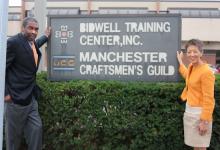 Bill Strickland and Jane Chu pose on either side of a vintage looking sign that reads Manchester Craftsmen's Guild