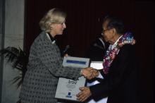 A woman with blond hair presents a large framed certificate to a man in glasses wearing a lei.