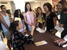 A woman sits behind a desk signing books with a crowd of people around her. 