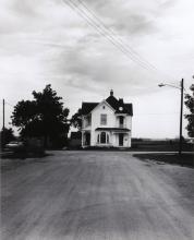 a two-story white wooden house seen at a distance down a road