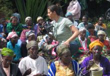  a white woman in Western dress holds a film camera while sitting amidst a crowd of Rwandan women 