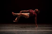 During a dance performance, Cameron McKinney, an African American man with locked hair, seems to float suspended in the air on his back, with just one hand touching the stage.