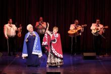 two older Latina women wearing embroidered capes sing while a mariachi band plays in the background