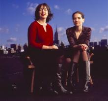 Two women seated on a rooftop open sky and clouds and the city skyline in the background.