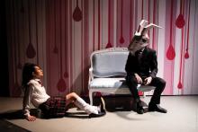 a girl in a school uniform sits on the floor looking up at a person sitting on a couch wearing an animal head