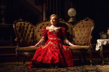 Marg Helgenberger in character as Regina Giddens on an early 1900s sofa wearing a red dress