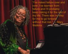 an older black woman with medium length locks and glasses sits at a black grand piano