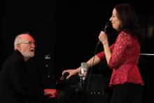 A man performs at the piano with a woman standing next to the piano singing.