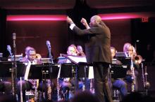 A man stands facing an orchestra with his arms raised