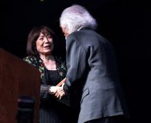 Japanese woman shaking hands with white-haired man in suit on stage. 