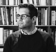 Portrait of man wearing dark glass frames in front of a bookcase. 