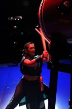 Japanese-American woman in costume banging on a taiko drum. 