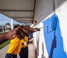 A hand in the foreground rolls blue paint on a cement block wall in the creation of a mural