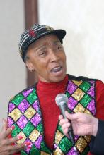 A woman wearing a red shirt and brightly sequined hat and vest speaks into a microphone.