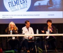 A woman, a man, and another woman holding a microphone and answering a question sit behind a long table with a film screen behind them