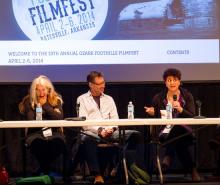 A woman, a man, and another woman holding a microphone and answering a question sit behind a long table with a film screen behind them