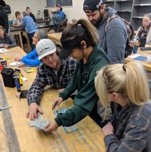 a group of students sitting at a long table making postcards