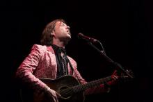 a white man with medium long hair in a patterned red jacket plays a guitar and sings into a microphone