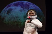 Boy in astronaut suit in front of photo of the moon. 