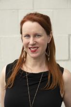 A woman with red hair stands against a white wall.