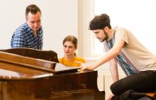 female student sits at piano with two men looking at sheet music with her