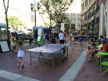 People on the sidewalk of a city doing hula hoop near a ping-pong table. 