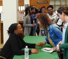 A woman sits behind a table shaking hands with a woman across the table who is talking to her.