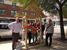 Two teens cut a ribbon on a newly installed arrow sculpture while two adult men stand smiling at the camera