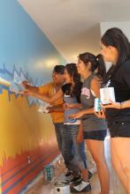 Four young Native American girls in casual clothing paint a mural in blue, yellow, and orange on an office wall